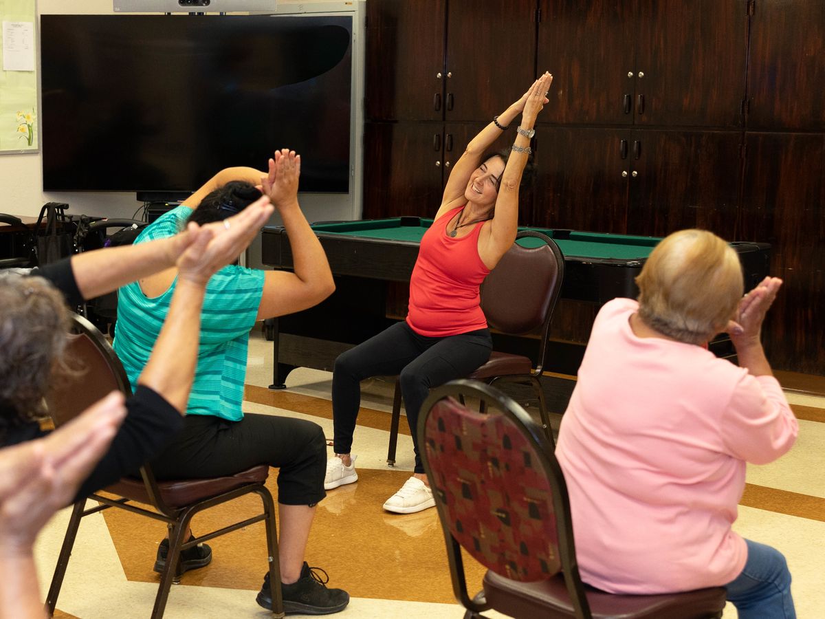 Nikki demonstrates a seated overhead stretch as participants mirror her.